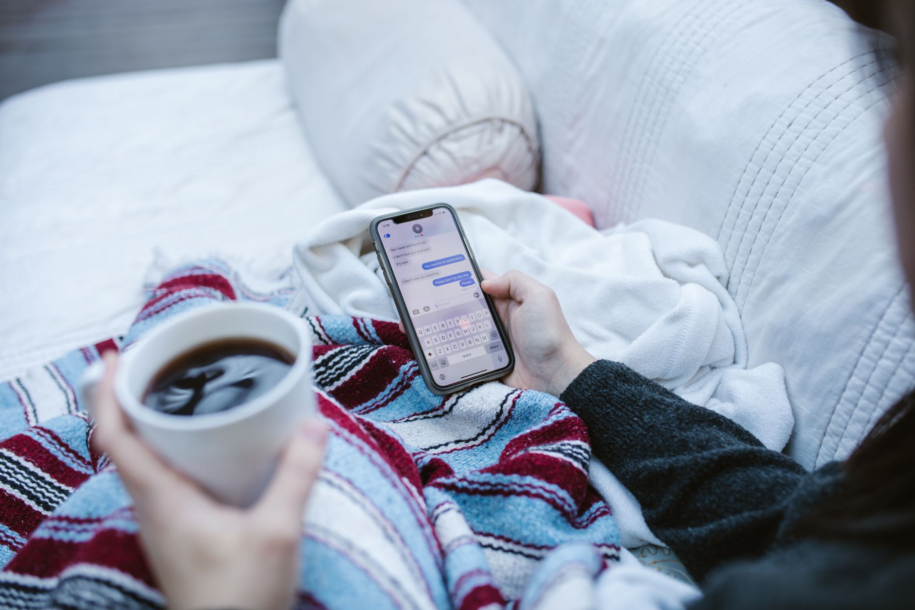 Personne sous une couverture rayée tenant un smartphone qui affiche une conversation par messages et une tasse de café noir.